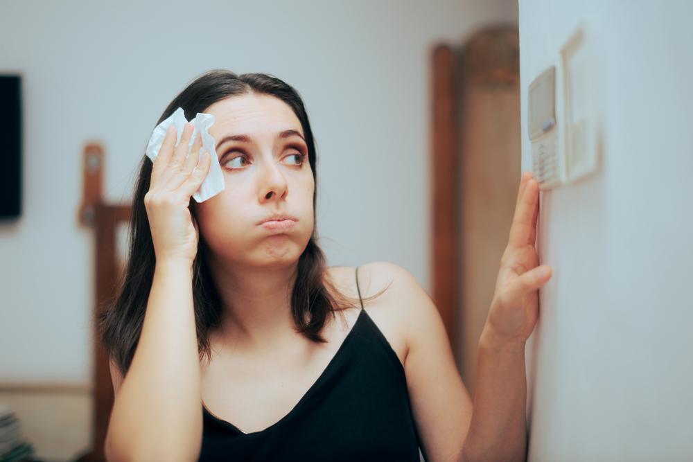 A woman looking at the thermostat while wiping sweat from her forehead because her AC is blowing hot warm air.